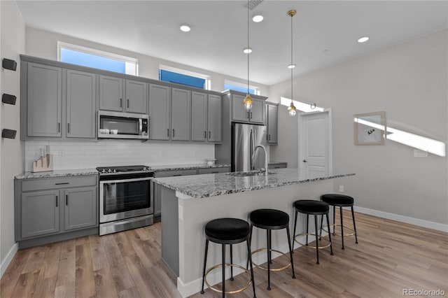 kitchen with stainless steel appliances, a kitchen island with sink, light stone countertops, and gray cabinetry