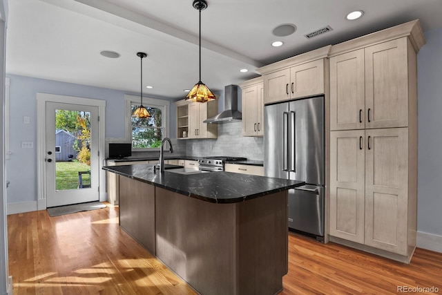 kitchen with a center island with sink, dark stone countertops, stainless steel appliances, wall chimney exhaust hood, and decorative light fixtures