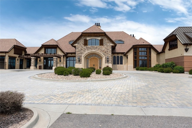 french country home featuring a tile roof, stone siding, stucco siding, a chimney, and curved driveway