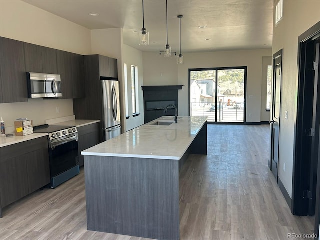 kitchen with sink, appliances with stainless steel finishes, a kitchen island with sink, decorative light fixtures, and light wood-type flooring