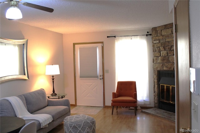 living room featuring light wood-style flooring, a stone fireplace, a ceiling fan, and a textured ceiling