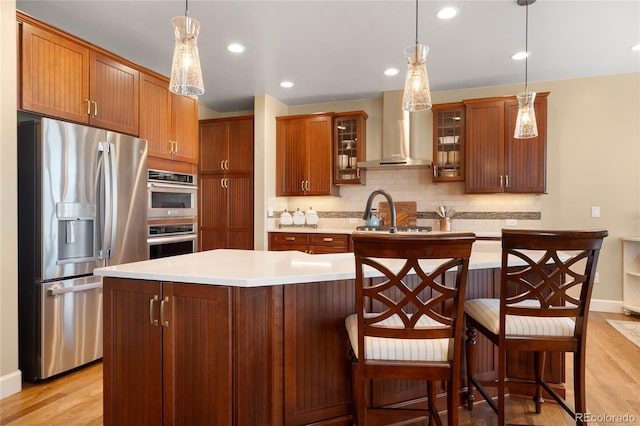 kitchen featuring a center island with sink, wall chimney exhaust hood, stainless steel appliances, and decorative light fixtures