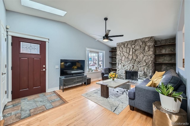 living room featuring a ceiling fan, wood finished floors, vaulted ceiling with skylight, a stone fireplace, and baseboards