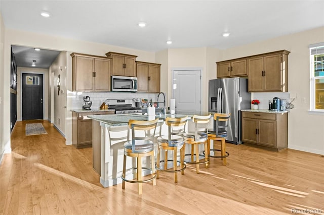 kitchen with an island with sink, a kitchen bar, light wood-type flooring, appliances with stainless steel finishes, and light stone counters