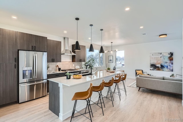 kitchen with a center island with sink, wall chimney exhaust hood, light countertops, appliances with stainless steel finishes, and a kitchen breakfast bar