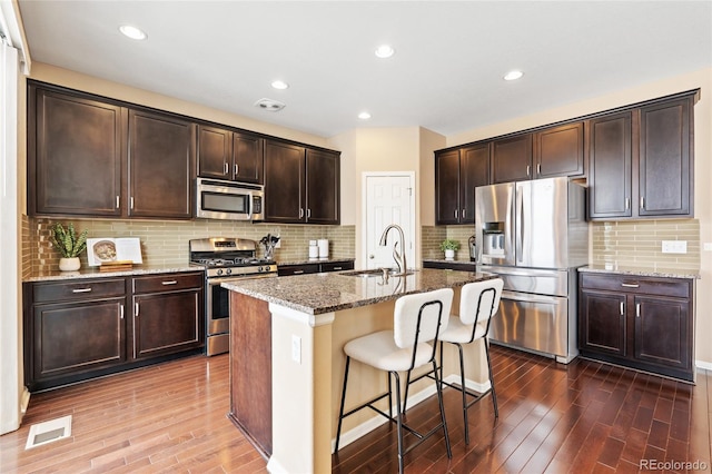 kitchen featuring dark wood-type flooring, sink, light stone counters, a center island with sink, and stainless steel appliances