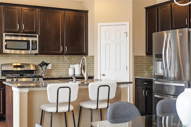 kitchen with stainless steel appliances, a breakfast bar area, a center island with sink, and dark stone countertops