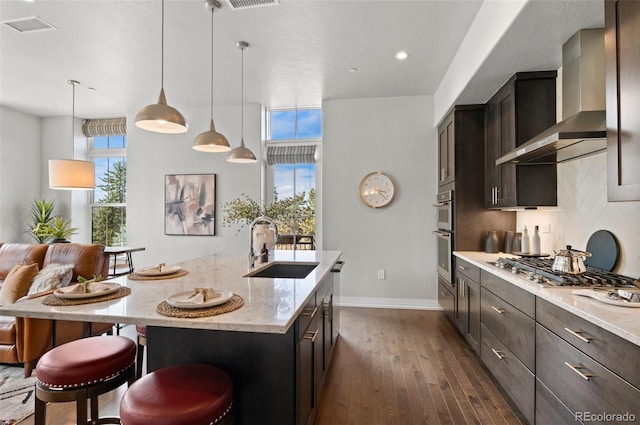 kitchen with wall chimney range hood, light stone countertops, decorative backsplash, appliances with stainless steel finishes, and a sink