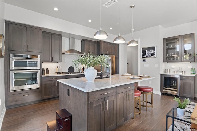 kitchen featuring a kitchen bar, visible vents, beverage cooler, stainless steel appliances, and wall chimney range hood