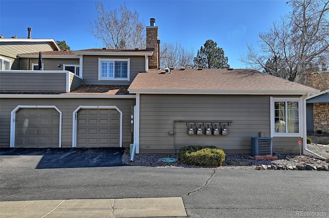 view of front of home with central AC unit, a garage, driveway, roof with shingles, and a chimney