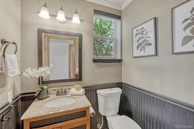 half bath featuring ornamental molding, a textured wall, vanity, toilet, and wainscoting
