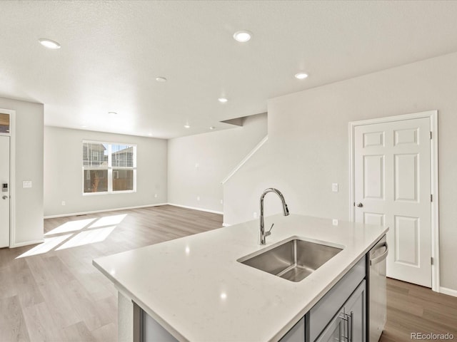 kitchen with sink, a kitchen island with sink, stainless steel dishwasher, light stone countertops, and light hardwood / wood-style flooring