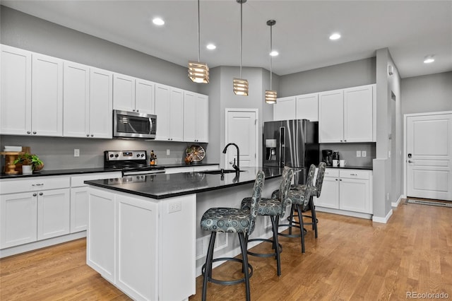 kitchen with a kitchen island with sink, stainless steel appliances, a sink, light wood-type flooring, and dark countertops