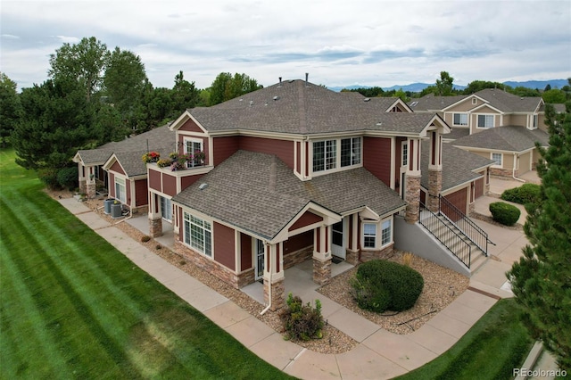 view of front facade featuring driveway, central AC unit, stone siding, roof with shingles, and a front yard