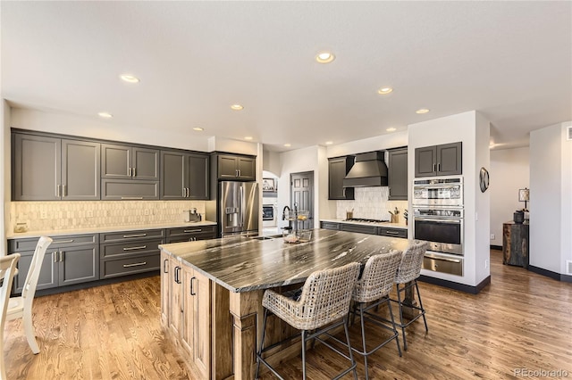 kitchen with a warming drawer, a center island with sink, a sink, appliances with stainless steel finishes, and wall chimney range hood