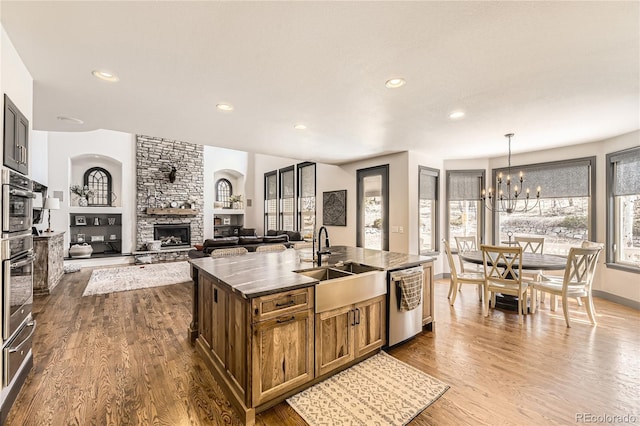 kitchen featuring a center island with sink, a stone fireplace, stainless steel dishwasher, dark wood-style floors, and a sink