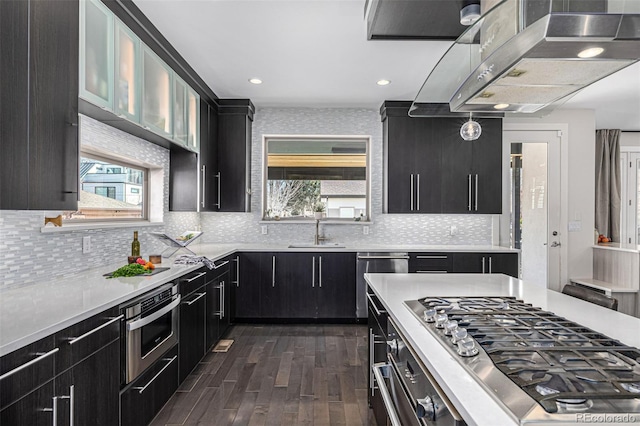 kitchen with a sink, wall chimney range hood, stainless steel appliances, light countertops, and dark cabinets