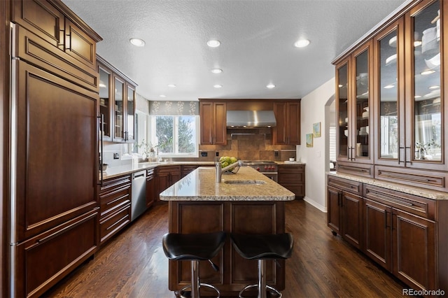 kitchen featuring an island with sink, dishwasher, dark hardwood / wood-style flooring, paneled refrigerator, and wall chimney exhaust hood