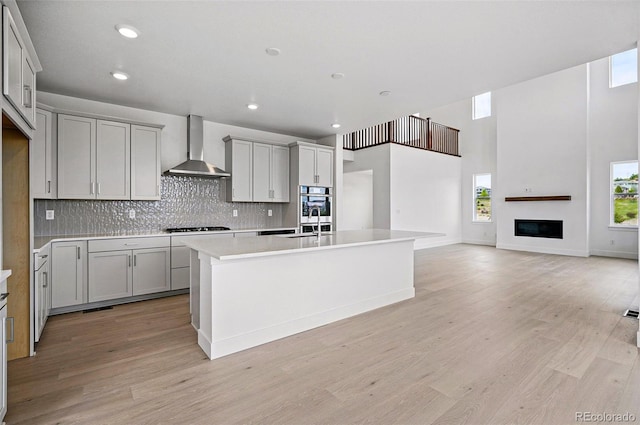 9382 Quaker St, Arvada, CO 80007 - kitchen featuring wall chimney range hood, gray cabinetry, light wood-style floors, an island with sink, and recessed lighting