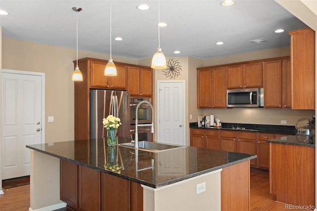 kitchen featuring decorative light fixtures, an island with sink, dark stone counters, stainless steel appliances, and light wood-type flooring