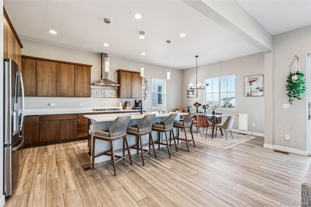 kitchen with light wood-style floors, light countertops, wall chimney range hood, appliances with stainless steel finishes, and an island with sink