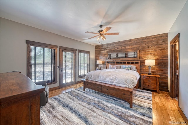 bedroom featuring wooden walls, ceiling fan, an accent wall, access to outside, and light wood-type flooring