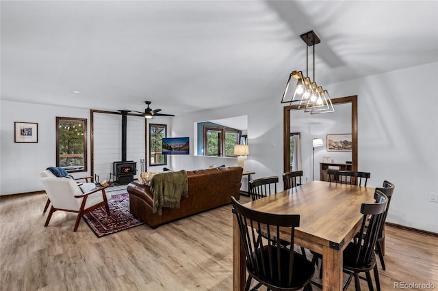 dining area with ceiling fan with notable chandelier, light wood-type flooring, a wealth of natural light, and a wood stove