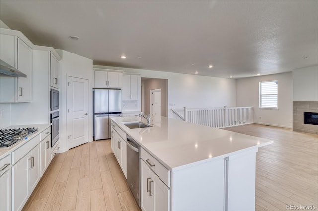 kitchen with appliances with stainless steel finishes, an island with sink, sink, white cabinets, and light wood-type flooring