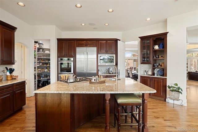 kitchen with a breakfast bar area, a kitchen island with sink, built in appliances, light stone counters, and light wood-type flooring