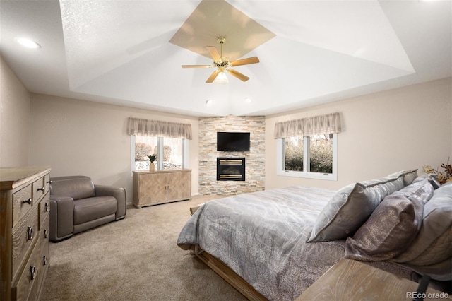 bedroom with a raised ceiling, carpet floors, a stone fireplace, and ceiling fan