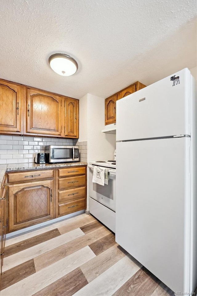 kitchen with white appliances, light wood-style flooring, under cabinet range hood, brown cabinets, and backsplash