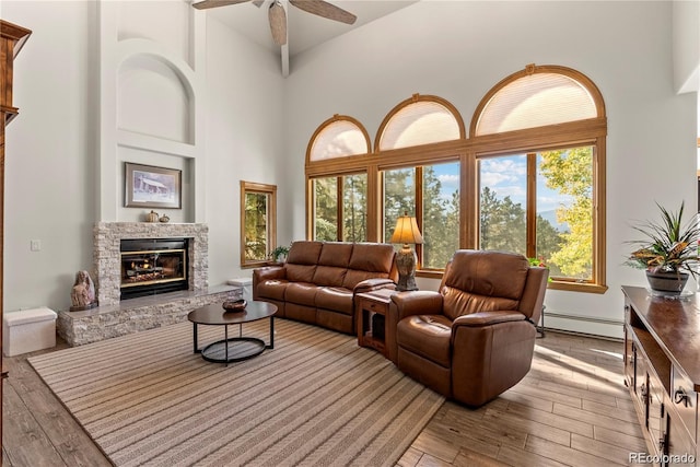 living area with a towering ceiling, a baseboard heating unit, wood finished floors, and a stone fireplace