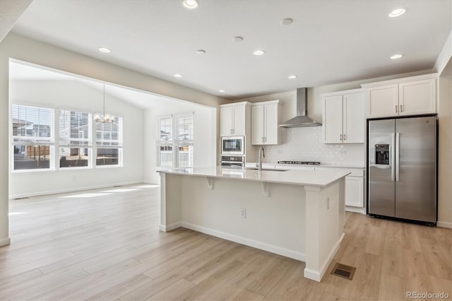 kitchen featuring sink, appliances with stainless steel finishes, wall chimney range hood, a kitchen island with sink, and white cabinets