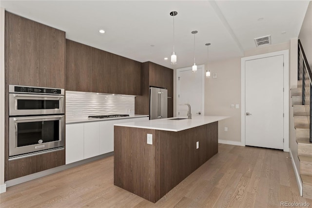 kitchen featuring white cabinetry, light wood-type flooring, an island with sink, stainless steel appliances, and hanging light fixtures