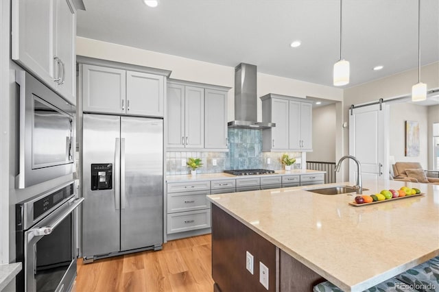 kitchen featuring wall chimney exhaust hood, a barn door, a sink, appliances with stainless steel finishes, and light wood-style flooring