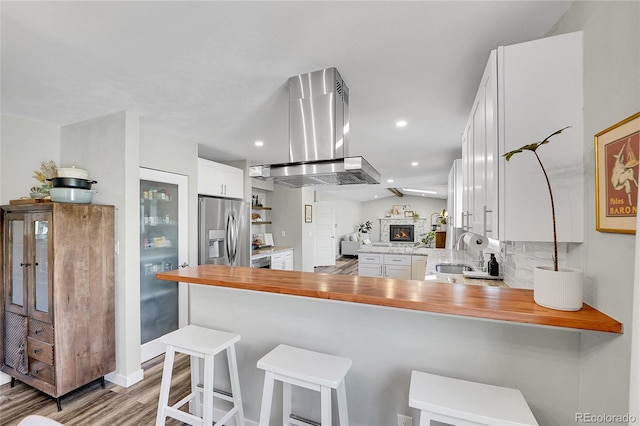 kitchen with a peninsula, a stone fireplace, white cabinets, stainless steel fridge, and island range hood