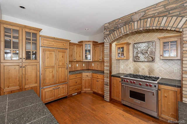 kitchen with tasteful backsplash, luxury range, brown cabinetry, glass insert cabinets, and dark wood-style floors