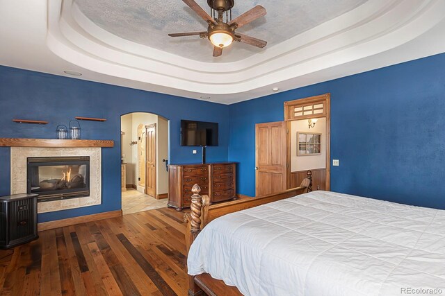 bedroom with a glass covered fireplace, a tray ceiling, hardwood / wood-style flooring, and arched walkways