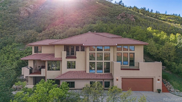 view of front of property featuring an attached garage, a wooded view, stucco siding, and a balcony