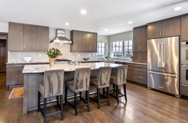 kitchen with wall chimney range hood, a kitchen island with sink, dark brown cabinets, and high end fridge