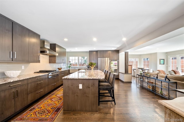 kitchen with a breakfast bar area, stainless steel appliances, open floor plan, wall chimney range hood, and a center island with sink