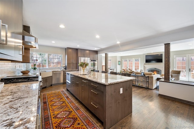 kitchen with appliances with stainless steel finishes, a center island with sink, dark brown cabinets, and light stone countertops