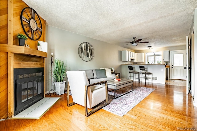 living room with a tiled fireplace, ceiling fan, a textured ceiling, and light wood-type flooring