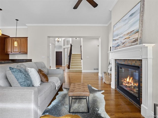 living area with stairs, a ceiling fan, wood finished floors, a tile fireplace, and ornamental molding