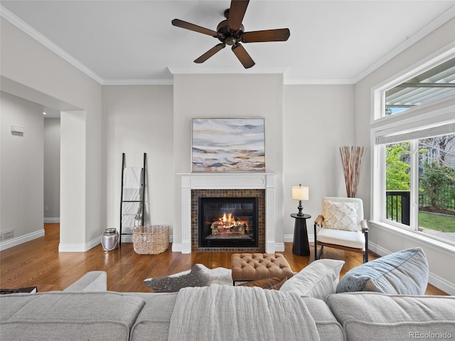 living room with a tiled fireplace, a ceiling fan, wood finished floors, visible vents, and ornamental molding