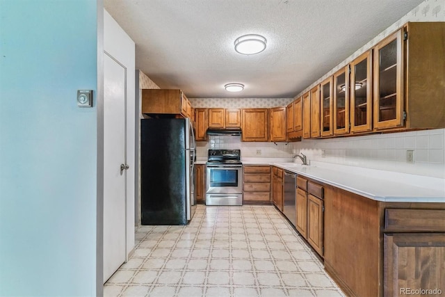 kitchen featuring stainless steel appliances, brown cabinetry, under cabinet range hood, light flooring, and glass insert cabinets