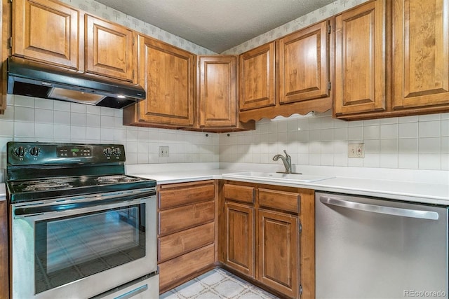 kitchen featuring appliances with stainless steel finishes, under cabinet range hood, a sink, brown cabinetry, and a textured ceiling