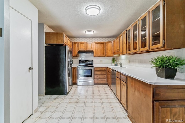 13626 E Bates Ave Unit 110, Aurora, CO 80014 - kitchen with appliances with stainless steel finishes, under cabinet range hood, brown cabinetry, a textured ceiling, and glass insert cabinets