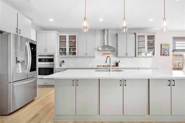 kitchen featuring decorative light fixtures, light countertops, appliances with stainless steel finishes, wall chimney range hood, and glass insert cabinets