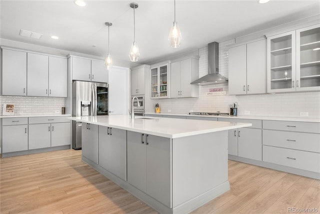 kitchen featuring glass insert cabinets, decorative light fixtures, a kitchen island with sink, light countertops, and wall chimney range hood